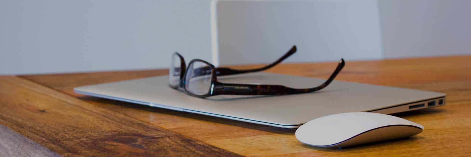 pair of spectacle glasses ontop of closed laptop with computer mouse on desk